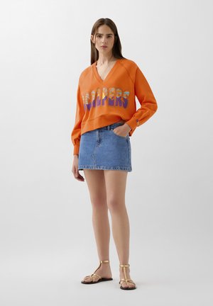 Felpa arancione con testo grafico, scollatura a V e maniche lunghe, abbinata a una gonna mini in denim e sandali golden a T.