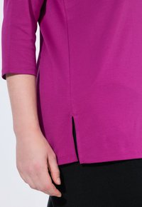 Fuchsia korte mouw tuniek met een zijsplit en een gladde textuur. Naaddetails en een ronde halslijn. Close-up van de stof en hand.