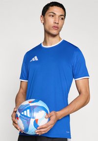 Camiseta deportiva azul con ribete blanco, que muestra el logo de Adidas. El modelo sostiene un balón de fútbol colorido con patrones en espiral.