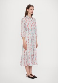 Cvečna midi obleka v svetlo modri barvi z rdečimi cvetovi, vertikalnimi črtami, gumbnim zapiranjem spredaj in vezanjem v pasu; nosi se z črnimi ravnimi čevlji.