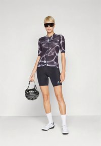 Personne portant une tenue de cyclisme noire et blanche, des lunettes de soleil et des chaussures de cyclisme blanches, tenant un casque de cyclisme noir par les sangles.
