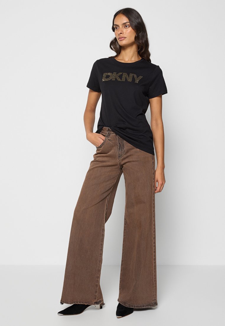 Czarna bawełniana koszulka z złotym logo, zestawiona z brązowymi szerokimi dżinsami. Modelka w czarnych botkach na obcasie, stojąca na neutralnym tle.