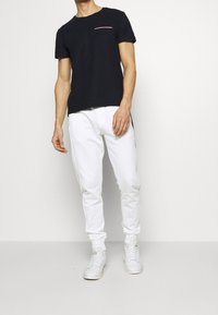 Schwarzes Baumwoll-T-Shirt mit einem rosa Taschenakzent, kombiniert mit weißen Jogginghosen. Beide Teile haben eine lässige Passform und glatte Texturen. Weiße Sneaker runden den Look ab.