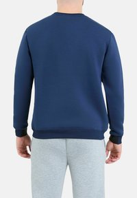 Granatowy sweter z miękkiego materiału, z okrągłym dekoltem, z prążkowanymi mankietami, noszony z jasnoszarymi spodniami dresowymi, gładkimi, bez wzorów czy akcentów.