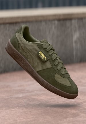 Olivgrüner Puma Palermo Sneaker aus Wildleder und strukturiertem Stoff, braune Sohle und ein Logo-Anhänger, der über gepflastertem Boden schwebt.