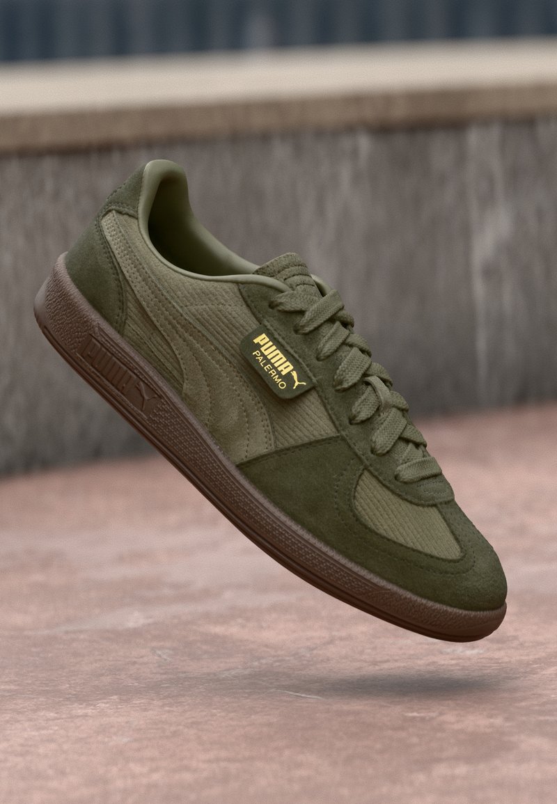 Basket Puma Palermo vert olive en suède et tissu texturé, semelle marron et étiquette logo flottant au-dessus d'un sol pavé.