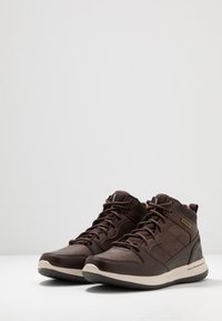 Zapatillas altas marrones hechas de cuero y textil, con puntera redondeada, collar acolchado, cordones y suela de goma con diseño texturizado.