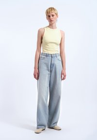 Un débardeur côtelé jaune clair et un jean large bleu clair. Le jean a un délavage et deux poches avant. Le mannequin porte des chaussures beige.