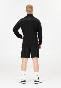 Man i svart långärmad träningströja, svarta shorts, vita ankelsockor och vita sneakers står med handen på höften vänd bort från kameran mot en vit bakgrund.