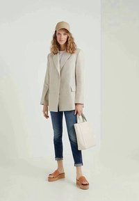 Blazer bege de dois botões, t-shirt cinza-claro, jeans escuros, sandálias plataforma castanhas e um boné bege. Segura uma bolsa tote branca.