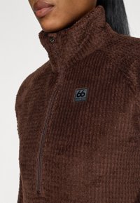 Pullover de fleece castanho com colarinho alto, fecho de zip e tecido texturizado. Apresenta um logotipo preto da 66° North no lado esquerdo.