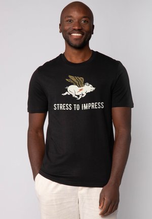 Schwarzes T-Shirt mit einem Grafikdesign eines fliegenden Schweins mit Flügeln und einem Blitz, der Text lautet "STRESS TO IMPRESS". Kurze Ärmel, runder Halsausschnitt.