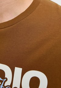 Brązowa koszulka bawełniana z okrągłym dekoltem oraz dużym białym nadrukiem z przodu. Miękka faktura z detalem w postaci ściągacza przy kołnierzu.