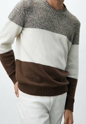 Man wearing a long-sleeve knit sweater with gray, white, and brown horizontal color blocks, paired with white pants.