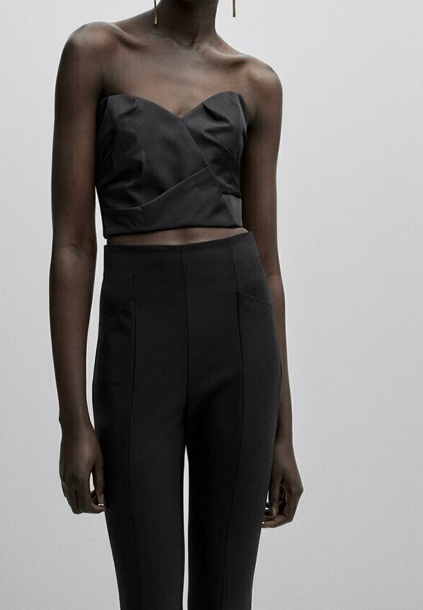 Femme portant un top court noir sans bretelles et un pantalon noir taille haute sur un fond gris clair uni.