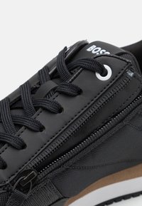 Zapatilla deportiva negra con parte superior texturizada, puntera redondeada, cierre de cordones y cremallera lateral. Detalles incluyen logo blanco y suela marrón.