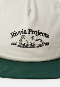 Casquette beige avec une visière verte. À l'avant, on trouve un texte brodé en noir "Rivvia Projects" ainsi qu'une illustration de serpent. Matière en coton.