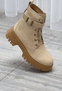Beige semskede ankelstøvler har et snørebånddesign, en sidestropp med metallspenne og en kraftig brun gummisåle med mønster.