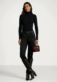 Sieviete melnā turtleneck blūzē, melnās kravas biksēs un augstās melnās zābakos, tur mazu sarkanu čehu somu pret vienkāršu fonu.