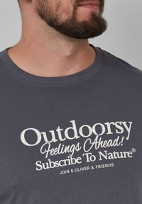 Donkergrijze t-shirt met korte mouwen met vetgedrukte witte tekst: "Avontuurlijke Gevoelens Vooruit! Abonneer Je Op Natuur." Ronde halslijn en zachte stof.