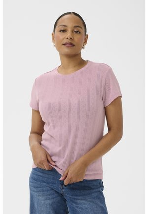 Femme aux cheveux tirés en arrière portant un haut texturé rose à manches courtes et un jean bleu sur un fond blanc uni.