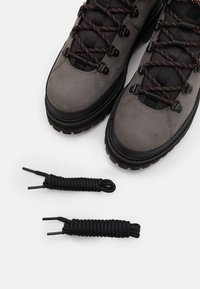 Harmaat vaelluskengät mustilla yksityiskohdilla ja oransseilla nauhoilla; rakenne on nahkaa, joka on teksturoitu; mukaan tulee myös kaksi ylimääräistä mustaa nauhaa solmuilla.