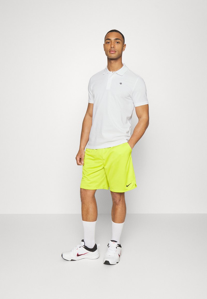 Hvit polo skjorte med en liten logo, parret med lyse neongule shorts. Synlige hvite sokker og svarte joggesko med røde detaljer.