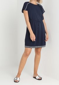 Robe bleu marine à manches courtes avec taille à cordon, ornée de broderies blanches le long des manches et de l'ourlet. Portée avec des sandales noires à enfiler.