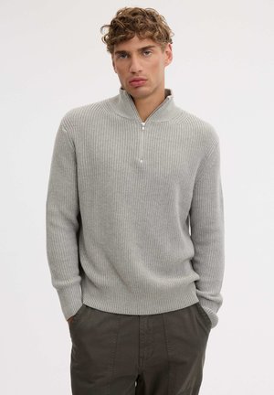 Un hombre luce un jersey gris claro de canalé con cremallera corta y unos pantalones oscuros. Está de pie con las manos en los bolsillos ante un fondo liso.