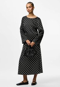 Vestido negro de manga larga con lunares blancos, silueta fluida, acompañado de un pequeño bolso negro y zapatos planos negros de puntera.