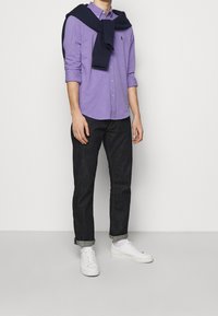 Fialová košile na knoflíky, námořnický svetr přehozený přes ramena, černé džíny, bílé tenisky, stojící na jednobarevném pozadí.