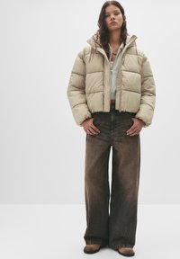 Giacca imbottita beige con taglio squadrato, lunghezza cropped e motivo trapuntato visibile. Abbinata a jeans marroni a gamba larga e calzature marroni.