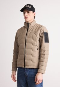 Beige vadderad jacka med hög krage, utrustad med ett diagonalt quiltat mönster, svart dragkedja och en ficka med dragkedja på vänster ärm.