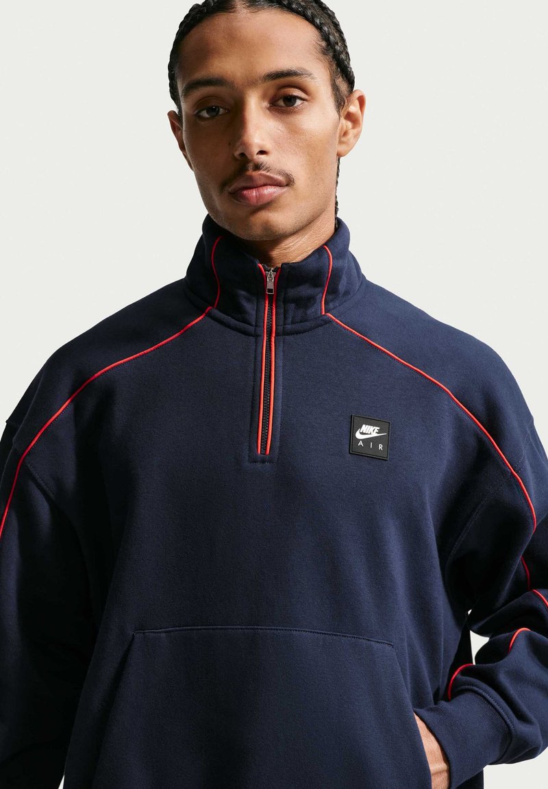 Jeune homme aux cheveux tressés portant un sweat-shirt Nike Air marine à fermeture éclair au quart, avec un liseré rouge, debout devant un fond uni.