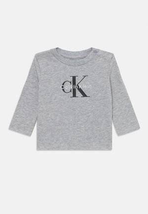 Chemise grise à manches longues en coton, arborant un grand logo 'CK' noir et le texte "Calvin Klein" en blanc, avec un bouton-pression à l'épaule.