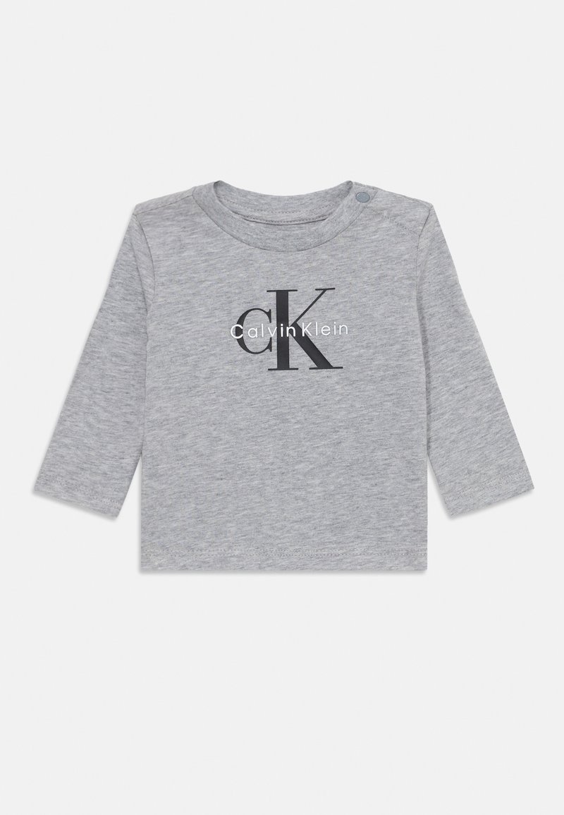 Grå langermet skjorte laget av bomull, med en stor svart 'CK'-logo og teksten "Calvin Klein" i hvitt, med en trykknapp på skulderen.