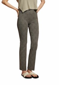 Vrouw in een grijs geruite slim-fit broek en beige kruisband sandalen, staand met handen op de heupen tegen een witte achtergrond.
