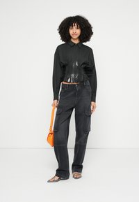 Maglia nera a maniche lunghe con una vita aderente e lucida, abbinata a pantaloni cargo scuri. La modella tiene una borsa arancione e indossa sandali argentati.