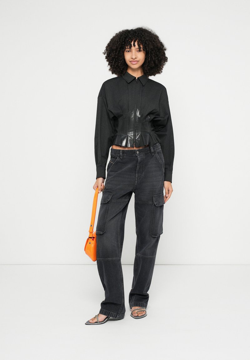 Schwarzes langärmliges Hemd mit tailliertem, glänzendem Bund, kombiniert mit dunklen Cargohosen. Das Modell hält eine orangefarbene Handtasche und trägt silberne Sandalen.