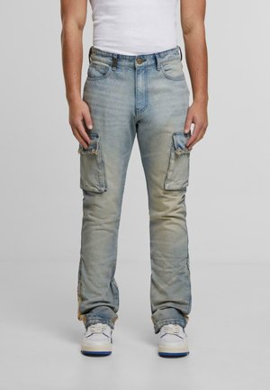 Man wearing faded blue denim cargo jeans with frayed knee pockets, white sneakers, and a white sleeveless shirt, standing against a plain backdrop.