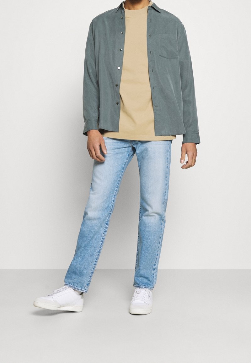 Camisa ligera de botones en verde sobre una camiseta beige, combinada con jeans rectos de color azul claro y zapatillas blancas.