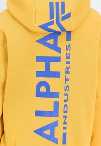 Sweat à capuche jaune avec un grand texte vertical bleu « Alpha Industries » imprimé dans le dos, s'étendant de la capuche jusqu'au bas du dos.