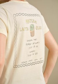 Světlé žluté bavlněné tričko s potiskem grafiky matcha latte a textem s otevíracími hodinami a údaji o lokalitě v černé a zelené barvě.