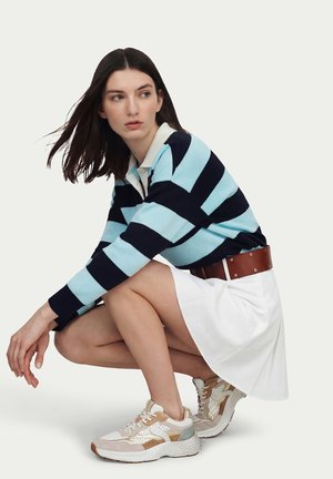 Donna accovacciata che indossa una maglia a maniche lunghe a righe blu e nere, gonna bianca con cintura marrone e scarpe da ginnastica beige e bianche su sfondo semplice.