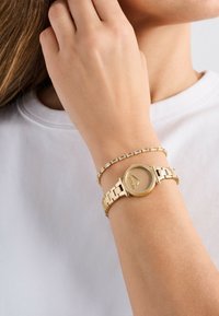 Žena nosící zlaté hodinky s texturovaným kulatým ciferníkem a zlatým náramkem s obdélníkovými krystaly na levém zápěstí.