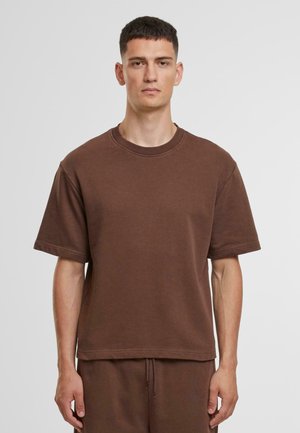 Egy férfi sima barna rövidujjú, kerek nyakú pólót és hozzáillő barna nadrágot visel, egy egyszínű világos háttér előtt áll.