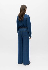 Femme aux longs cheveux bruns portant une chemise en denim bleu foncé ample et un jean taille haute à jambes larges, debout de dos sur un fond blanc uni.