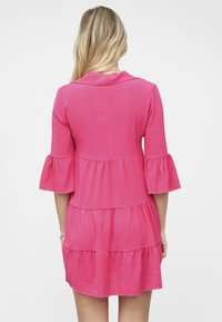 Vaaleanpunainen, kolmen neljänneksen hiha mekko, jossa on kauluskaulus ja kerroksellinen muotoilu. Valmistettu kevyestä, teksturoidusta kankaasta, jossa on liehuvat hihat.