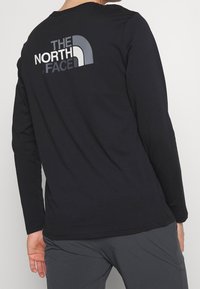 Schwarzes Langarmshirt aus weichem Stoff, mit einem großen grauen und weißen Logo-Design auf dem Rücken. Rundhalsauschnitt und lässige Passform.