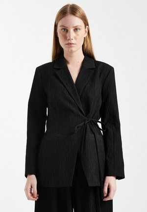 Donna con capelli rossi lisci che indossa un blazer nero strutturato con cintura in vita, rivolta frontalmente su uno sfondo bianco semplice.
